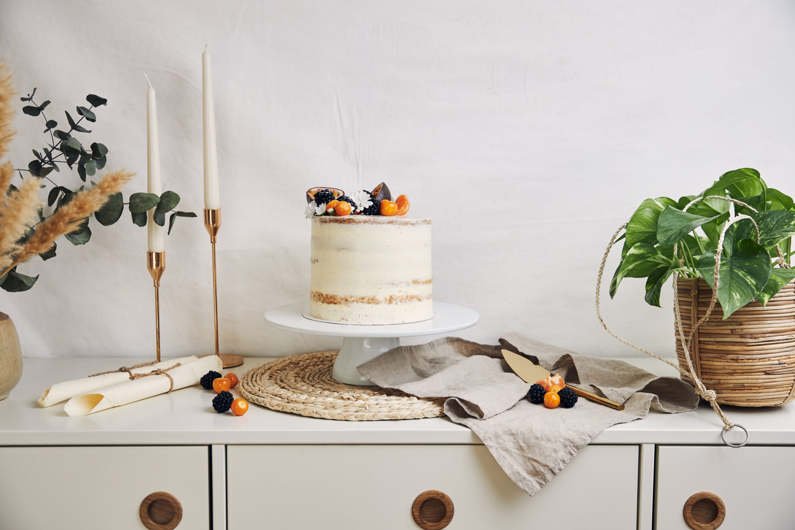 A white cake with berries and passionfruits next to plants and candles behind a white background
